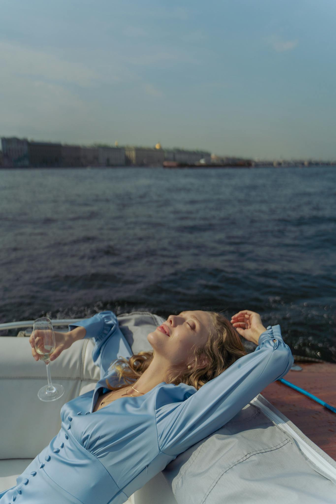 A woman in a blue dress relaxes on a boat holding a champagne glass.